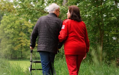 Ein älterer Mann mit Rollator spaziert in Begleitung einer Rotkreuz Mitarbeiterin auf einem Feldweg.
