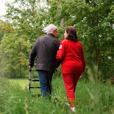 Ein älterer Mann mit Rollator spaziert in Begleitung einer Rotkreuz Mitarbeiterin auf einem Feldweg.