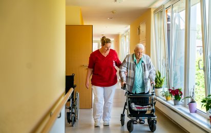 Eine Pflege-Mitarbeiterin begleitet eine Bewohnerin eines Seniorenwohnhauses bei einem Spaziergang