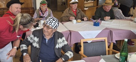 Im Vordergund spricht eine Pflegemitarbeiterin mit einem Bewohner, der auf seinem Kopf eine Sträflingskappe mit der Nummer 283 trägt. Im Hintergrund sitzen weitere Bewohnerinnen an einem Tisch.