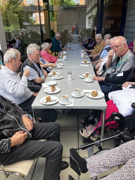 Die Bewohnerinnen und Bewohner des Seniorenwohnhauses sitzen auf der Terrasse an einem Tisch mit Kaffee und Kuchen