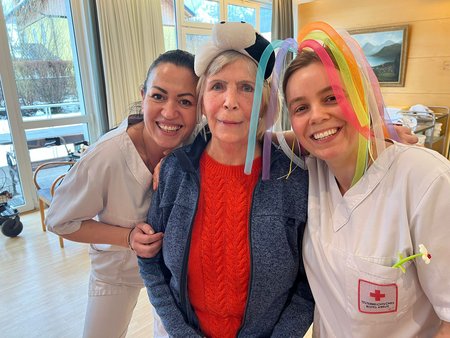 Zwei Pflegemitarbeiterinnen lachen gemeinsam mit einer in der Mitte stehenden Bewohnerin in die Kamera. Die rechte Pflegemitarbeiterin trägt einen Hut aus bunten Luftballonen.