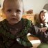 im Vordergrund krabbelt ein Kleinkind, im Hintergrund sieht man die Mutter, die am Boden sitzt und das Kind beobachtet