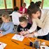 Kinder malen an einem Tisch sitzend und werden dabei von einer Frau angeleitet