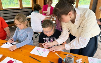 Kinder malen an einem Tisch sitzend und werden dabei von einer Frau angeleitet