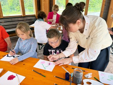 Kinder malen an einem Tisch sitzend und werden dabei von einer Frau angeleitet