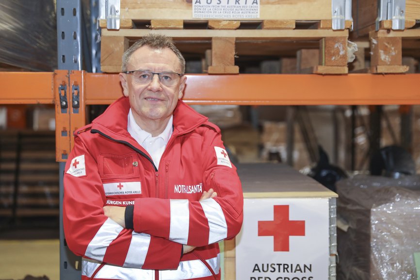 Ein älterer Mann in Rotkreuz Unfiform steht in vor Lagerregalen im Katastrophenhilfslager