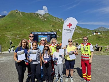 Erste-Hilfe-Kursteilnehmer:innen mit Rettungssanitäter:innen vor eine Rotkreuz-Fahne