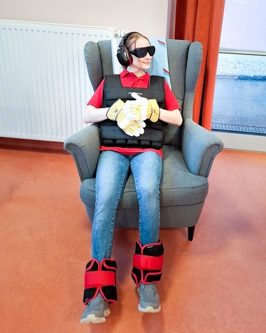 Eine junge Frau sitzt mit Gewichtsweste, Spezialbrille, Schutzhandschuhen, Schallschutzkopfhörern und Gewichtsmanschetten um den Knöcheln in einem Sessel.
