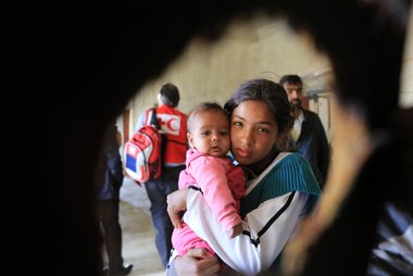 © IFRC/Ibrahim Malla Eine junge Frau mit einem Kleinkind im Arm schaut in die Kamera
