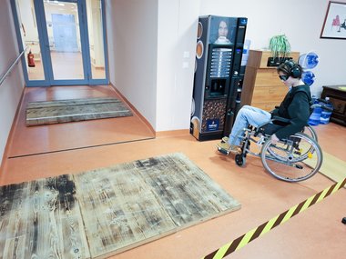 Eine Person mit Spezialbrille und Schallschutzkopfhörern fährt in einem Rollstuhl sitzend über einen Testparcours.