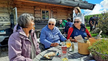 Drei Personen sitzen an einem Tisch auf einer Alm und essen