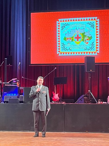 Martin Huber begrüßt in Rotkreuz-Uniform mit einem Mikrofon in der Hand die Gäste des Rotkreuz-Balles, im Hintergrund ist eine Bühne mit einer Leinwand und einer Abbildung der Fahne der Freiwilligen Kolonne Salzburg zu sehen.