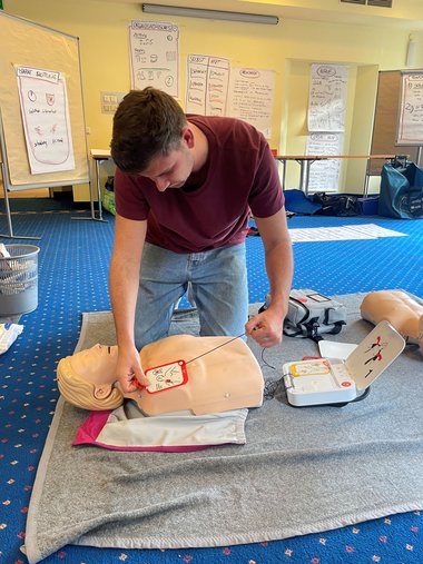Ein Mann kniet neben einer Übungspuppe und befestigt die Defibrillator-Pads an ihr.
