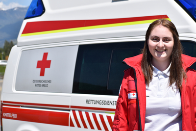 Sanitäterin Chiara steht vor einem Rettungswagen und trägt die markante rote Uniformjacke.