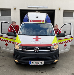 Vater Gerhard und Sohn Stefan stehen hinter den geöffneten Türen eines Rettungswagens. Man sieht den Wagen und die beiden frontal von vorne.