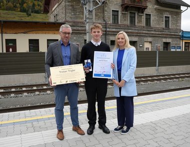 Drei Personen stehen an einem Bahnsteig und blicken in die Kamera. Eine Person hält eine Tafel in der Hand, eine andere ein Zertifikat.
