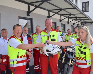 Mitarbeiter:innen vom Roten Kreuz Lungau stehen gemeinsam vor dem neuen Einsatz-Motorrad mit Helm und Schlüssel in der Hand