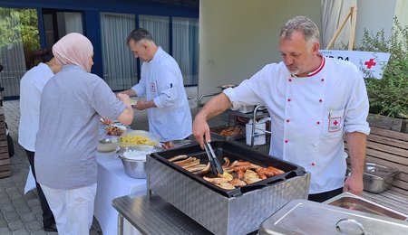 Zwei Köche bereiten Essen am Griller vor bzw. richten Salat auf einem Teller an, während zwei Frauen sich etwas zu essen holen