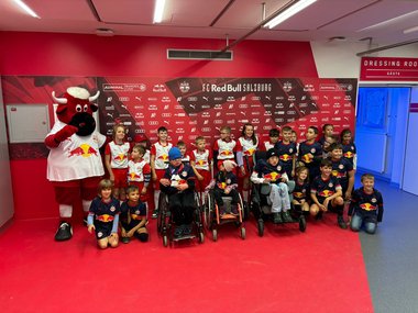 Gruppenfoto mehrerer Kinder in weiß-roten oder blauen Trikots des FC-Red-Bull-Salzburgs. Links neben der Gruppe steht das Maskottchen des Vereins. Im Hintergrund eine Sponsorenwand mit roter Grundfarbe.