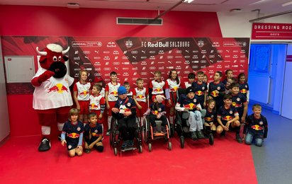Gruppenfoto mehrerer Kinder in weiß-roten oder blauen Trikots des FC-Red-Bull-Salzburgs. Links neben der Gruppe steht das Maskottchen des Vereins. Im Hintergrund eine Sponsorenwand mit roter Grundfarbe.