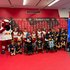 Gruppenfoto mehrerer Kinder in weiß-roten oder blauen Trikots des FC-Red-Bull-Salzburgs. Links neben der Gruppe steht das Maskottchen des Vereins. Im Hintergrund eine Sponsorenwand mit roter Grundfarbe.