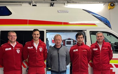 ein Mann wird von je 2 Rettungssanitäter flankiert, sie stehen vor einem Rettungsauto