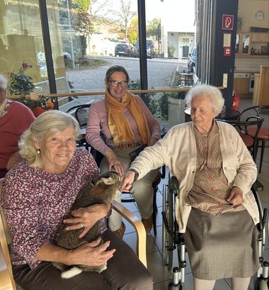 Eine ältere Dame sitzt und hält einen Hasen in Arm, eine zweite Dame streckt ihren Arm zu dem Hasen auf. Dahinter sitzt eine dritte Frau, die dabei zusieht.
