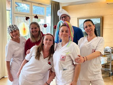 Gruppenfoto der Pflegemitarbeiterinnen. Eine von ihnen trägt einen bunten Hut aus Luftballonen, eine andere eine Sträflingsmütze mit der Zahl 283. Zwei der Mitarbeiterinnen tragen einen Haarreifen mit auf Federn befestigten Fußbällen beziehungsweise Käfern. Im Hintergrund ist ein als Schlump verkleideter Mitarbeiter zu sehen.