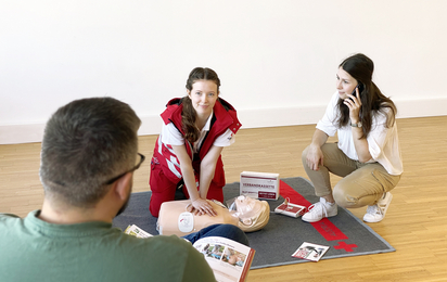Erste-Hilfe-Kurs Eine Erste-Hilfe-Lehrbeauftragte sitzt am Boden und zeigt eine Reanimation vor, während eine Person von hinten zuschaut und eine andere mit dem Handy telefoniert.