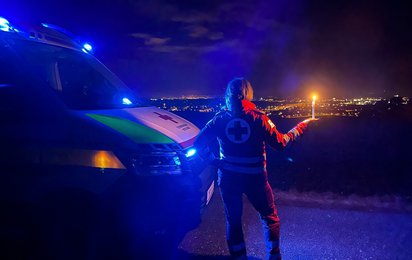 Eine Rotkreuz-Mitarbeiterin steht mit einer Kerze vor einem Rettungsauto, es ist dunkel und sie blickt auf eine beleuchtete Stadt hinunter.