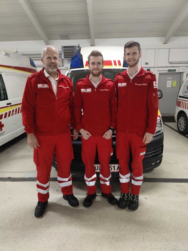 Vater Klaus mit seinen beiden Söhnen Leo und Felix, alle drei in Rotkreuz-Uniform, abgebildet vor einem SEW in der Rettungsgarage.
