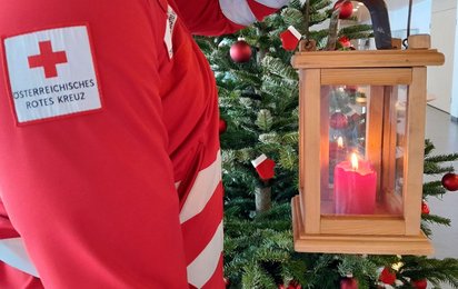 Eine hölzerne Laterne mit einer brennenden roten Kerze, umgeben von einem geschmückten Weihnachtsbaum und Dekorationen in rot und grün.