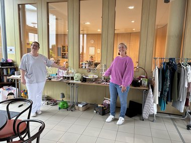 Die Hausleitung und eine Mitarbeiterin des Seniorenwohnhauses Obertrum präsentieren den Flohmarkttisch, der mit diversen Materialien und Gegenständen gefüllt ist.