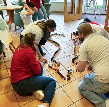 Mehrere Teilnehmerinnen sitzen oder knien am Boden und stellen gemeinsam Dominosteine auf.