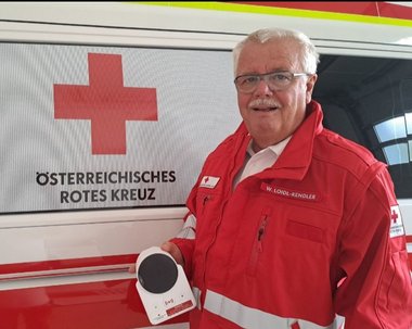 Wolfgang Loidl-Kendler freiwillig als Rufhilfe-Monteur, steht mit einem Rufhilfe Gerät vor einem Rotkreuz-Auto