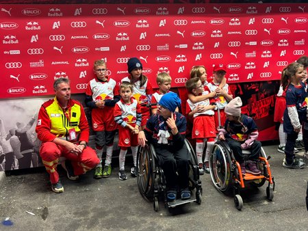Eine Gruppe mehrerer Kinder in weiß-roten und blauen Trikots des FC Red Bull Salzburgs, links daneben kniet ein Mann in Rotkreuz-Uniform mit gelber Weste.