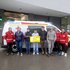 Eine Gruppe von Menschen steht vor einem Rettungswagen. Eine Person hält einen großen Scheck in Händen.