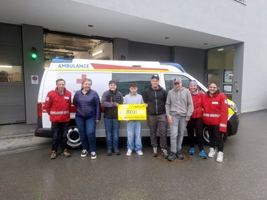 Eine Gruppe von Menschen steht vor einem Rettungswagen. Eine Person hält einen großen Scheck in Händen.