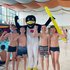 Vier Kinder in Badehosen stehen gemeinsam mit einem Menschen in einem Pinguinkostüm neben einem Schwimmbecken