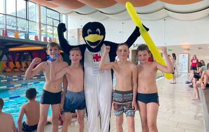 Vier Kinder in Badehosen stehen gemeinsam mit einem Menschen in einem Pinguinkostüm neben einem Schwimmbecken