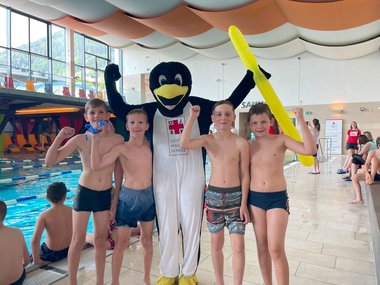 Vier Kinder in Badehosen stehen gemeinsam mit einem Menschen in einem Pinguinkostüm neben einem Schwimmbecken