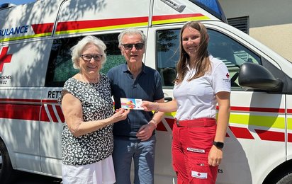 Eine Rotkreuz-Mitarbeiterin überreicht eine Hitzebuddy-Freecard älternen Menschn vor einem Rettungsauto.