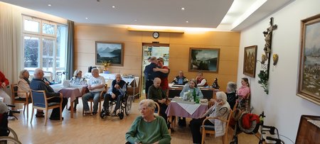 Das ganze Haus feiert beim Geburtstag mit. Zu sehen sind die Senior:innen sitzend in einem großen Raum. 