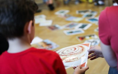 Zwei Kinder betrachten in einem Raum eine Bildkarte, während weitere Karten verstreut am Boden liegen. Die Szene zeigt eine Gruppensituation in Innenraumumgebung.