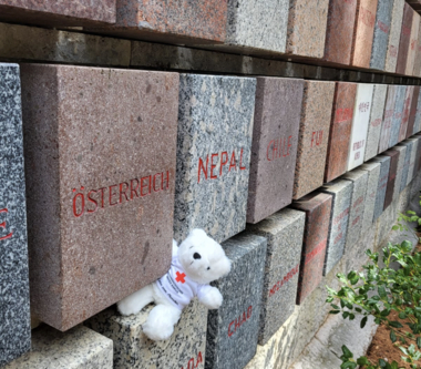 Erklärbär Bärli befindet sich an einer berühmten Mauer in Solferino und blickt dabei in die Kamera. Die Mauer besteht aus Steinen, in die alle Nationen der Welt, in der die Rotkreuz- und Rothalbmondbewegung vertreten ist, graviert sind.