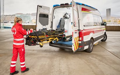 Eine Rettungssanitäterin steht hinten am Auto und zeigt die voll elektische Fahrtrage des Rettungswagens.