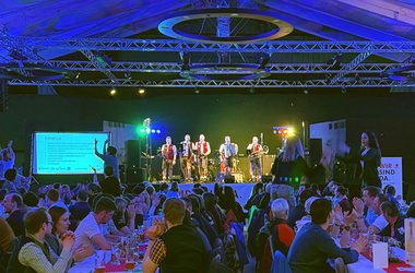 Aufnahme der Abschlussfeier der Bundesskimeisterschaft. In der Bildmitte ist auf der Bühne die Musikgruppe Die Alpenpiraten zu sehen. Im Vordergrund sitzen Menschen entlang langer Tische, manche klatschen mit der Musik mit.