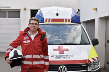 Maximilian Zarfl steht in Rotkreuz-Uniform vor einem Rettungswagen und hält dabei einen Schutzhelm unter dem Arm. An seiner linken Brust ist ein Funkgerät sichtbar.