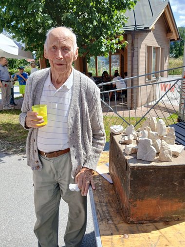 Ein älterer Herr steht mit einem Becher in der Hand neben einem Tisch mit Specksteinen